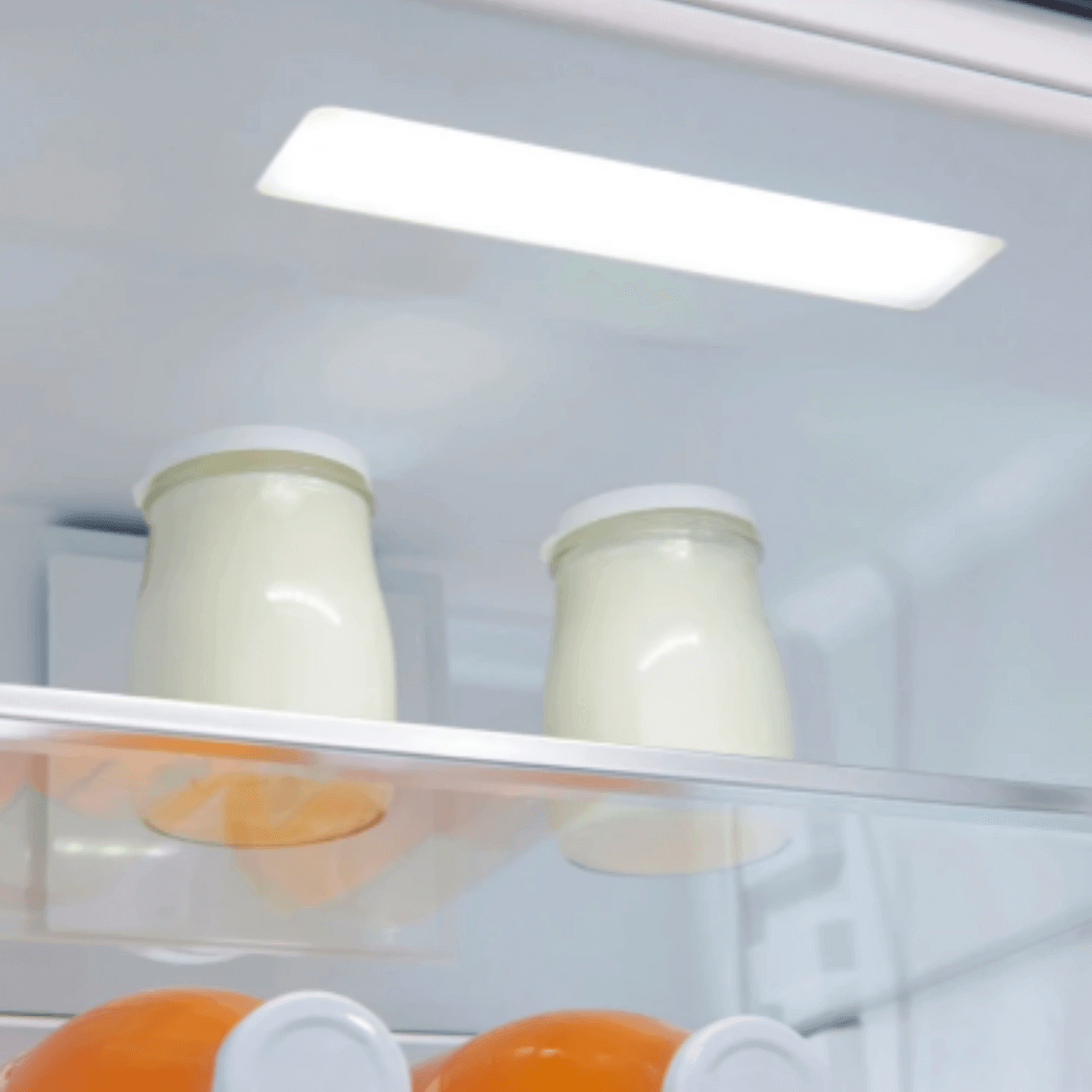 Interior do refrigerador com frascos de iogurte e laranjas na prateleira.