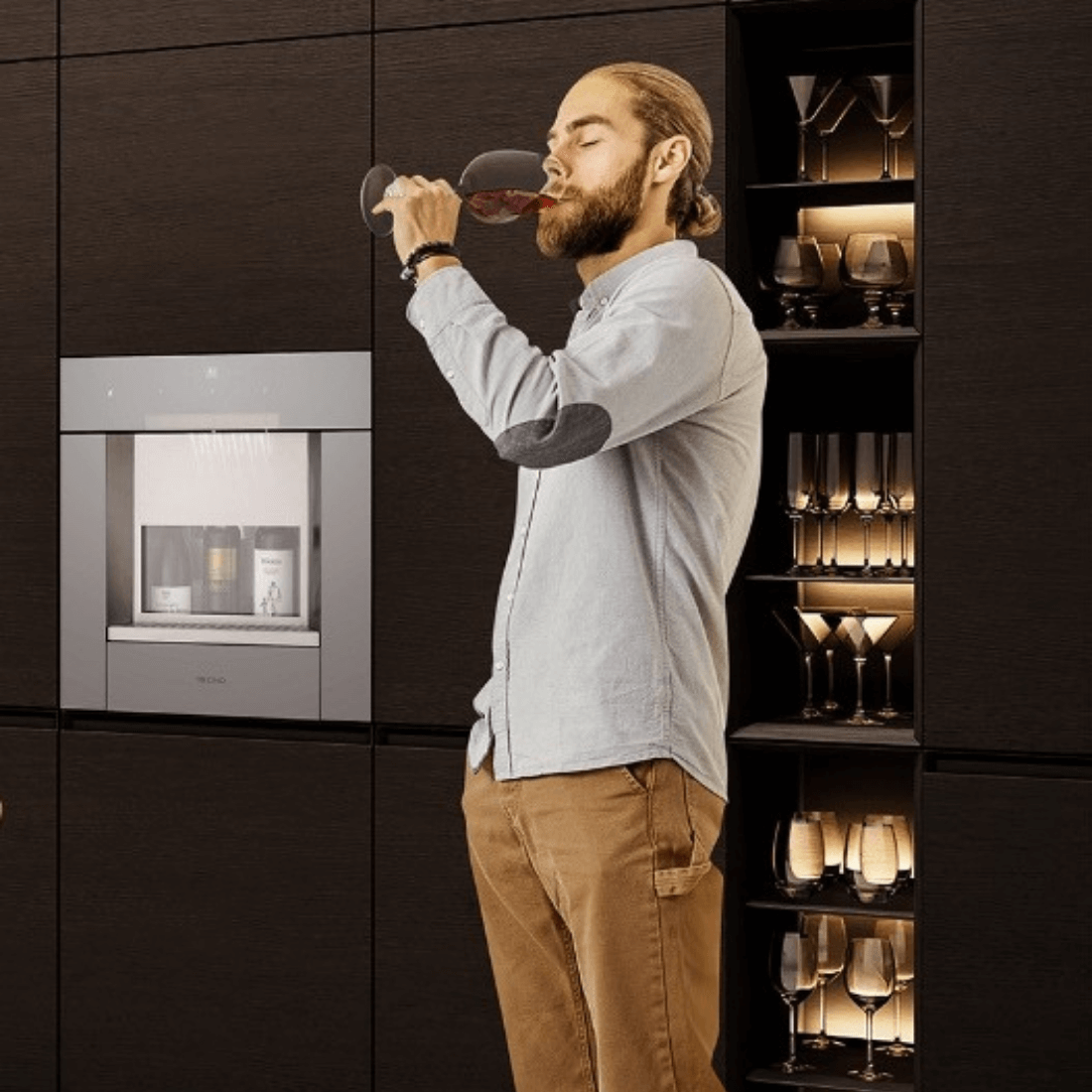 Homem degustando vinho ao lado de um dispenser de bebidas e taças em um armário elegante.