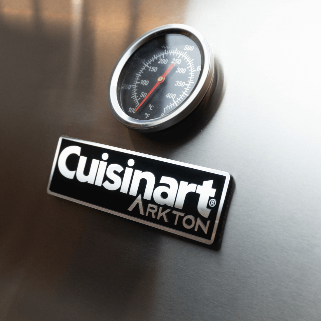 Cuisinart Arkton grill logo with temperature gauge on stainless steel surface.