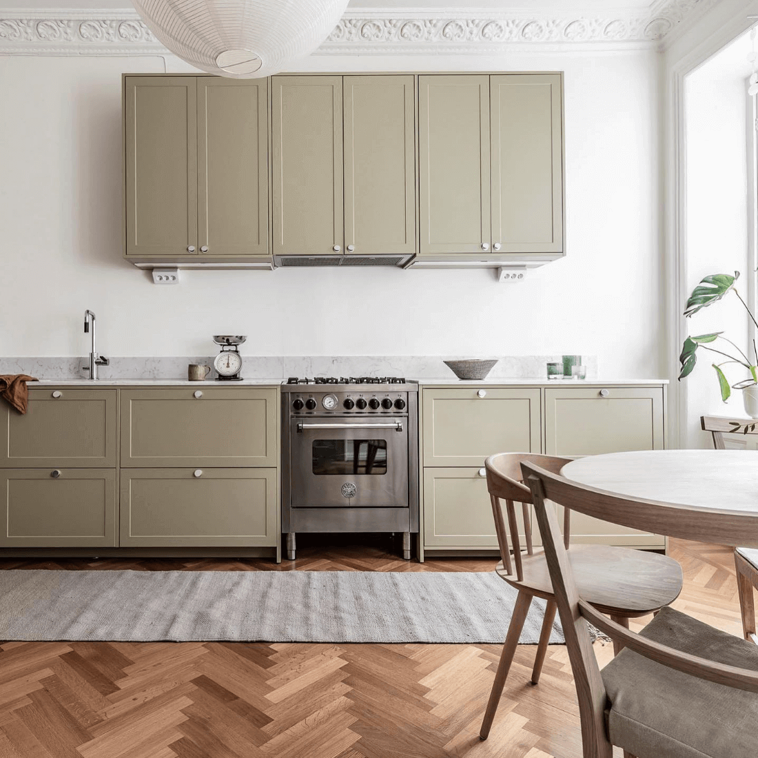 Cozinha moderna com armários verdes, fogão inox e mesa de madeira, decorada com plantas e estrutura minimalista.