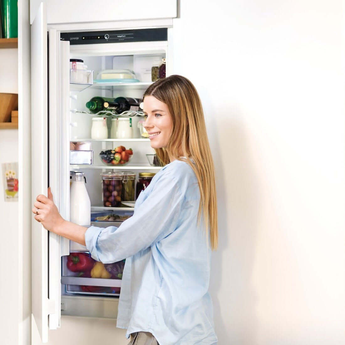 Mulher sorrindo enquanto abre o refrigerador Gorenje Bottom Freezer embutido na cozinha.
