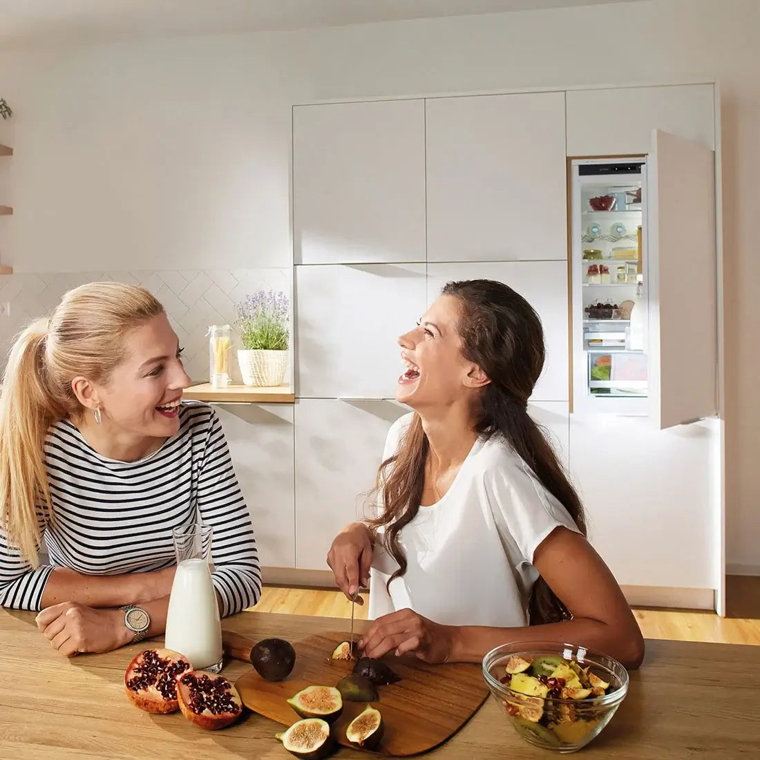 Duas mulheres sorrindo na cozinha, ao lado de um refrigerador embutido, preparando frutas frescas.