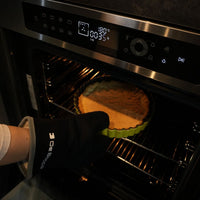 Forno Debacco Montreal Elétrico 73 Litros Vidro Preto 60cm mostrando torta sendo retirada com luva, porta de vidro e painel touch em inox