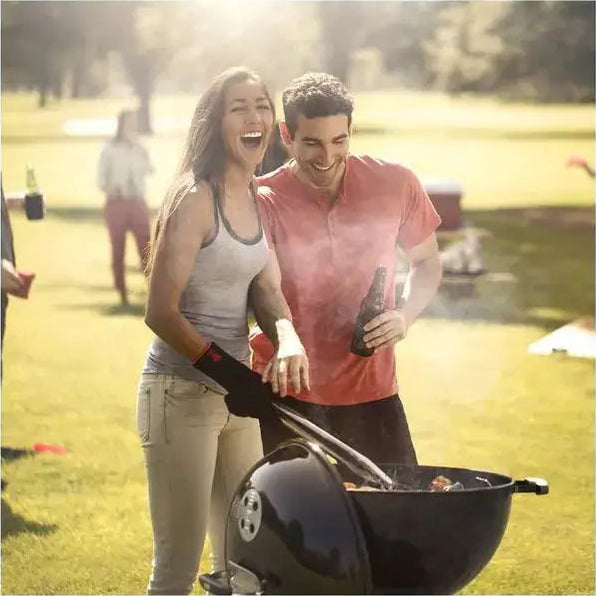 Churrasqueira a Carvão Weber Original Kettle: casal feliz grelhando em parque com churrasqueira preta, tampa, pegador e fumaça no convívio ao ar livre