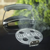 Weber Churrasqueira a Carvão Weber Original Kettle mostrada com tampa preta, puxador ergonômico e disco perfurado soltando fumaça