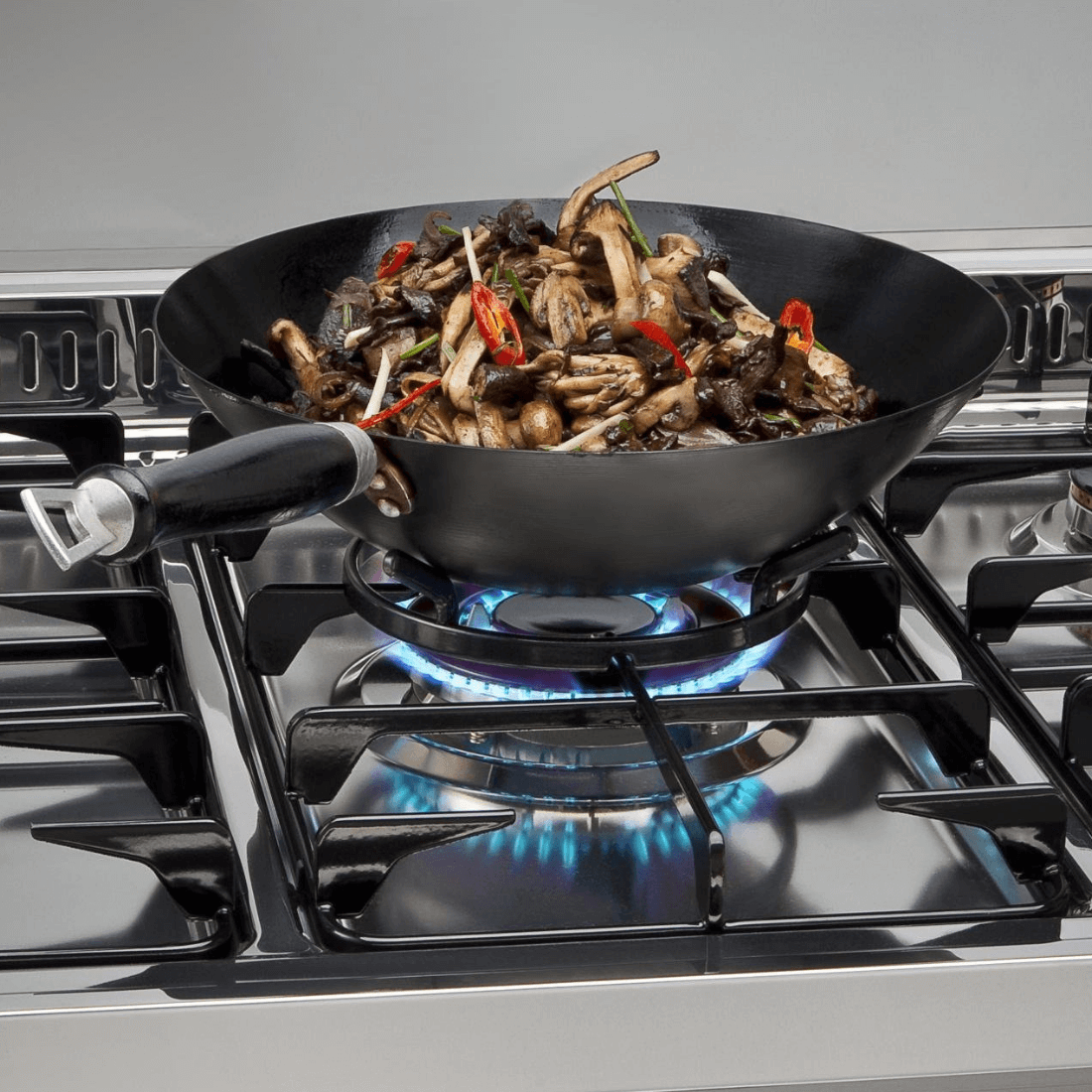 Wok com cogumelos e pimentas cozinhando em fogão Lofra, com chamas azuis visíveis.