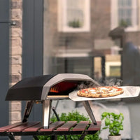 Forno de Pizza Portátil a Gás Ooni Koda 12 com pizza saindo quente e fumegante sob luz natural.