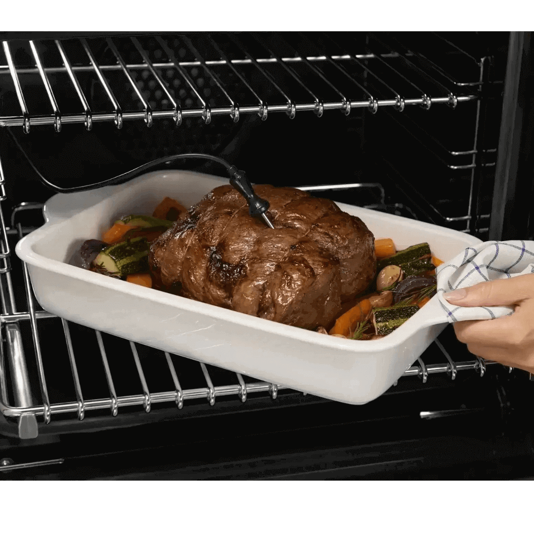 Assadeira com carne assada e legumes no forno, mão segurando toalha na borda.